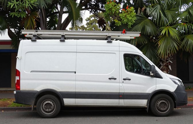 A white tradesman's van parked on a leafy Australian street with ladders on its roof rack